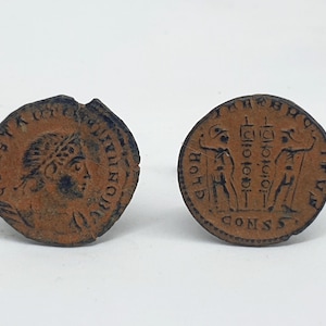 Op de afbeelding: Twee antieke bronzen munten. Eén munt toont een profiel van een persoon met een lauwerkrans en tekst. De andere munt heeft twee figuren en tekst. De munten liggen op een witte achtergrond.