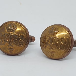 Puede incluir: Un par de gemelos dorados con un diseño de escudo de armas real. El escudo presenta una corona y las letras "G.R."