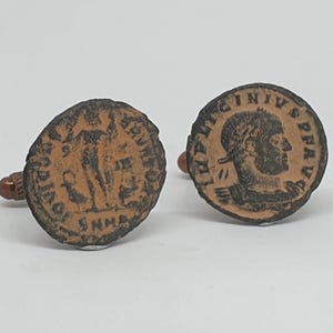 Op de afbeelding: Paar antieke manchetknopen met ronde, bronskleurige munten. Eén manchetknop toont een profiel van een persoon, de andere een staande figuur. De manchetknopen hebben een bruine, afgeronde paal.