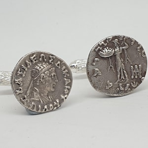Op de afbeelding: Zilveren manchetknopen met antieke muntontwerpen. Eén manchetknop toont een profiel van een persoon met een kroon en Griekse tekst. De andere toont een figuur met een object en extra symbolen.