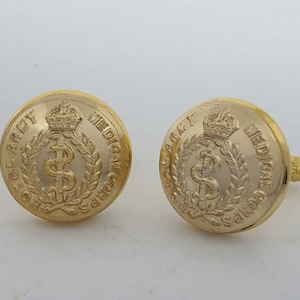 Puede incluir: Un par de gemelos dorados. Cada gemelo presenta las palabras "ROYAL ARMY MEDICAL CORPS" alrededor del borde, rodeando un símbolo de caduceo y una corona. Los gemelos tienen un acabado brillante.
