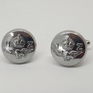 Puede incluir: Un par de gemelos plateados. Cada gemelo presenta un emblema en relieve con una corona, un águila y las letras "N" y "Z". Los gemelos son redondos y tienen un acabado metálico brillante.