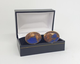 Peut inclure: Une paire de boutons de manchette ovales arborant un motif marbré bleu profond et doré métallisé, présentés dans un coffret de présentation rectangulaire noir à charnières avec une doublure blanche et un fin liseré doré sur le couvercle.