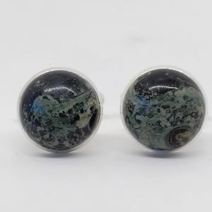 May include: A pair of round stud earrings. Each earring features a dark, marbled stone with shades of blue, green, and brown set in a silver-colored metal. The earrings are displayed against a white background.