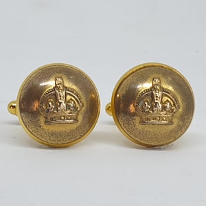 Puede incluir: Un par de gemelos dorados. Cada gemelo es redondo y tiene un diseño de corona en relieve en el centro. Los gemelos tienen un aspecto vintage ligeramente deslustrado.