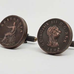 Puede incluir: Un par de gemelos con monedas antiguas de color cobre. Una moneda representa a Britannia, una figura femenina que representa a Gran Bretaña, sentada en una roca sosteniendo un tridente y un escudo. La otra moneda presenta un retrato de perfil del rey Jorge III, con la inscripción "GEORGIUS III D.G. REX" y la fecha "1805".