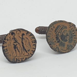 Op de afbeelding: Een paar manchetknopen met antieke Romeinse munten. De manchetknopen zijn gemaakt van metaal en hebben een bruine patina. De ene manchetknoop heeft een profiel van een Romeinse keizer, terwijl de andere een Romeinse soldaat toont.