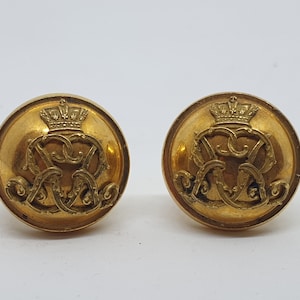 Puede incluir: Dos botones dorados antiguos. Cada botón presenta una corona sobre un monograma ornamentado. Los botones son redondos con un diseño ligeramente en relieve y un acabado brillante. Probablemente para ropa.