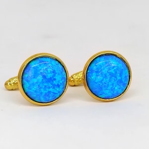 May include: A pair of gold-toned cufflinks with round, vibrant blue opal stones. The opals have a swirling, textured appearance, set in a gold bezel. The cufflinks are displayed on a white surface.