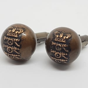 May include: A pair of cufflinks with a gold crown and a design of three circles with a crown on top. The cufflinks are made of a dark brown metal.