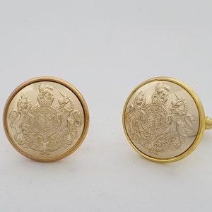 Może przedstawiać: Dwa złote spinki do mankietów z wypukłym wzorem królewskiego herbu. Spinki do mankietów są okrągłe i mają błyszczące wykończenie.