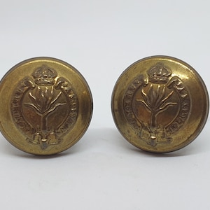 Puede incluir: Dos botones antiguos de latón con un emblema detallado que presenta una corona, una planta estilizada y texto circundante. Los botones son redondos y tienen un aspecto ligeramente envejecido.