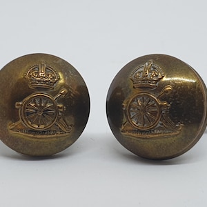 Puede incluir: Un par de gemelos dorados con un diseño de corona y rueda de cañón. Los gemelos están hechos de metal y tienen un acabado pulido.