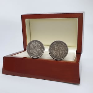 Puede incluir: Un par de gemelos de plata en una caja de terciopelo rojo. Los gemelos son redondos y presentan un diseño de perfil de hombre en un lado y un escudo de armas en el otro.