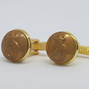 Puede incluir: Un par de gemelos de oro con un diseño circular marrón que presenta una corona y un símbolo. Los gemelos están conectados por una cadena de oro.