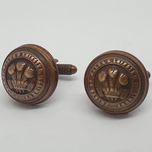 May include: A pair of bronze cufflinks featuring a raised design of three stylized feathers with a crown above them. The cufflinks are surrounded by a circular inscription that reads "Wales Elizabeth I" in a serif font.
