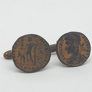 Op de afbeelding: Paar antieke bronzen manchetknopen met gedetailleerde Romeinse muntontwerpen. Eén manchetknop toont een profielportret, de andere een staande figuur. De manchetknopen hebben een getextureerde, bruine afwerking tegen een witte achtergrond.