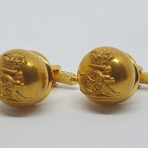 May include: A pair of gold-colored cufflinks. Each cufflink features a round, domed design with a detailed emblem of a crown and a cannon. The connecting bars have a textured, ridged pattern. The cufflinks are set against a white background.