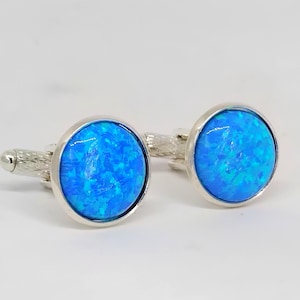 May include: A pair of silver cufflinks featuring round, bright blue opal stones. The opals have a swirling, textured appearance. The cufflinks have a textured silver stem and a rounded, polished silver backing.