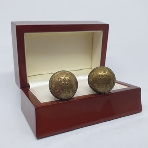 May include: A pair of antique brass cufflinks with engraved text, presented in a polished wooden box with a cream-coloured interior. The cufflinks are round and feature the text "THE DOG OR CARAB".