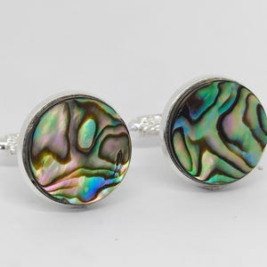 May include: A pair of round cufflinks with iridescent abalone shell inlays. The cufflinks have a silver-colored metal border and a textured post. The abalone shell displays a mix of green, blue, and purple hues.