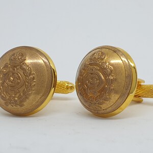 May include: A pair of gold cufflinks with a detailed engraved design featuring a crown and a wreath.