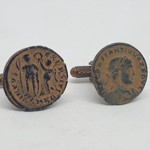 Op de afbeelding: Een paar manchetknopen met antieke Romeinse munten. De manchetknopen zijn gemaakt van metaal en hebben een bruine afwerking. De ene manchetknoop heeft een profiel van een Romeinse keizer, terwijl de andere twee figuren naast elkaar laat zien.