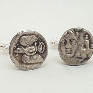 Op de afbeelding: Paar zilverkleurige manchetknopen met gedetailleerde antieke muntontwerpen. Eén manchetknop toont een zwaan, de andere een gezicht. De manchetknopen hebben een getextureerde, zilverkleurige pin.