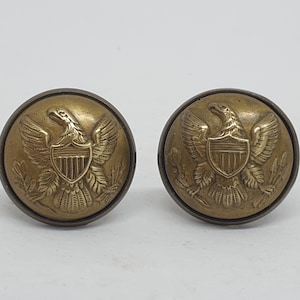 May include: Two gold-toned metal buttons with an eagle and shield design. The eagle is holding a shield with vertical stripes. The buttons are round and have a raised design.
