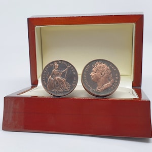 Puede incluir: Un par de gemelos en una caja de madera. Los gemelos están hechos de antiguos peniques británicos, uno con el perfil del rey Jorge III y el otro con Britannia. Los peniques están fechados en 1822.