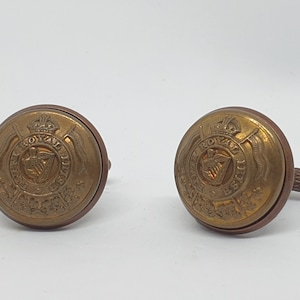 Puede incluir: Dos gemelos antiguos de latón. Cada gemelo presenta un diseño circular con un emblema, texto y un diseño de corona. Los gemelos tienen un aspecto deslustrado y envejecido, lo que sugiere que son antiguos. El texto dice "Royal Irish Fusiliers".