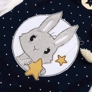 May include: A navy blue fabric with a white circle containing a gray bunny holding a yellow star. The background is covered in small white stars.