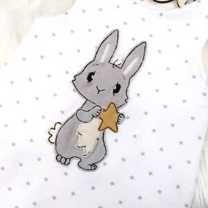 May include: A white onesie with a gray bunny holding a gold star. The onesie has a gray star pattern.