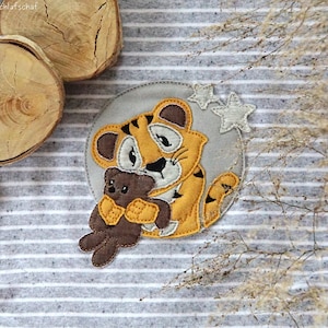 Könnte beinhalten: Ein braunes und oranges Tiger-Appliké, das einen braunen Teddybär hält. Das Appliké befindet sich auf einem grauen und weißen gestreiften Stoffhintergrund mit zwei weißen Sternen.