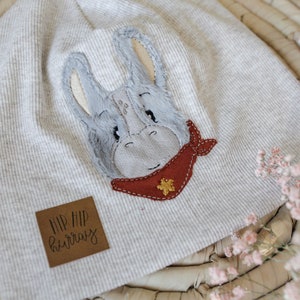 Könnte beinhalten: Eine graue gestrickte Baby-Mütze mit einem gestickten grauen und weißen Esel, der ein rotes Bandana mit einem goldenen Stern trägt. Die Mütze hat ein braunes Leder-Etikett mit der Aufschrift "Hip Hip Hooray".