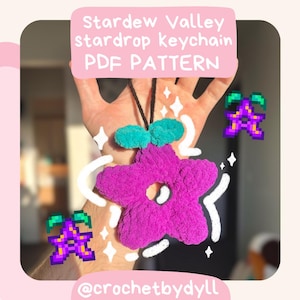 May include: Crocheted Stardew Valley stardrop keychain pattern. The keychain is purple with a green stem and leaves. The image includes the text "Stardew Valley stardrop keychain PDF PATTERN" and "@crochetbydyll".