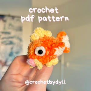May include: A handmade orange and yellow crochet fish with a white belly and black eye. The text "crochet pdf pattern" is in the upper portion of the image. The text "@crochetbydll" is in the lower portion of the image.