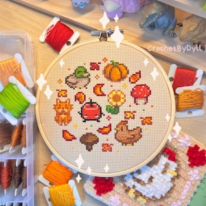 May include: A close-up of a completed cross-stitch embroidery hoop with a fall-themed design. The design features a variety of autumnal elements, including a pumpkin, leaves, mushrooms, a sunflower, a ladybug, a squirrel, and a chicken. The hoop is surrounded by colorful embroidery floss.