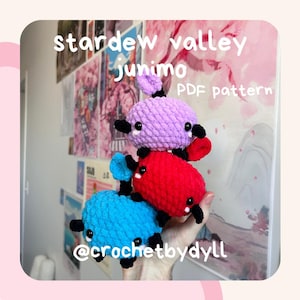 May include: Three crocheted Stardew Valley Junimo plushies stacked on top of each other. The top Junimo is purple, the middle Junimo is red, and the bottom Junimo is blue. The Junimos have black eyes and mouths. The image includes the text "stardew valley junimo PDF pattern" and "@crochetbydyll".