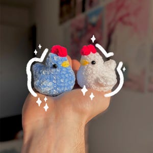 May include: Two handmade crochet chickens. One is blue with a red comb and yellow beak, the other is white with a red comb and yellow beak. The chickens are small and fluffy, perfect for a child's toy or home decor.