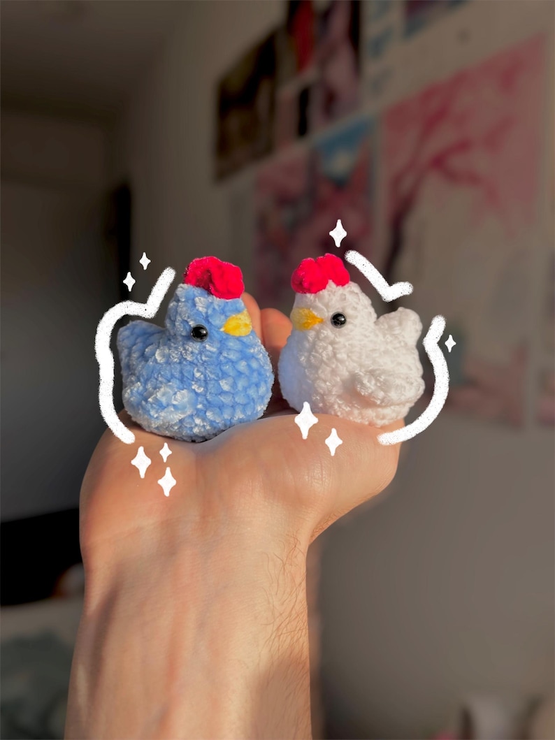 May include: Two handmade crochet chickens. One is blue with a red comb and yellow beak, the other is white with a red comb and yellow beak. The chickens are small and fluffy, perfect for a child's toy or home decor.