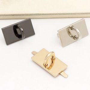 Könnte beinhalten: Drei rechteckige Metallteile für Handtaschenhardware mit einem Ring in der Mitte. Die Hardware ist in Schwarz, Silber und Gold dargestellt.