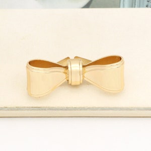 May include: A gold-coloured metal bow tie. The bow tie is a simple design with a smooth, polished finish.