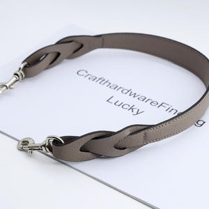 May include: A braided grey leather handbag strap with silver-coloured metal clasps. The strap is curved and rests on a white surface with the text "CrafthardwareFine Lucky". Designed for handbags.