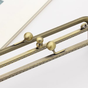 May include: A close-up of a gold-toned metal frame with a decorative pattern. The frame has multiple holes and is designed to be used as a purse or bag closure. The frame has three round metal knobs that can be used to adjust the length of the frame.