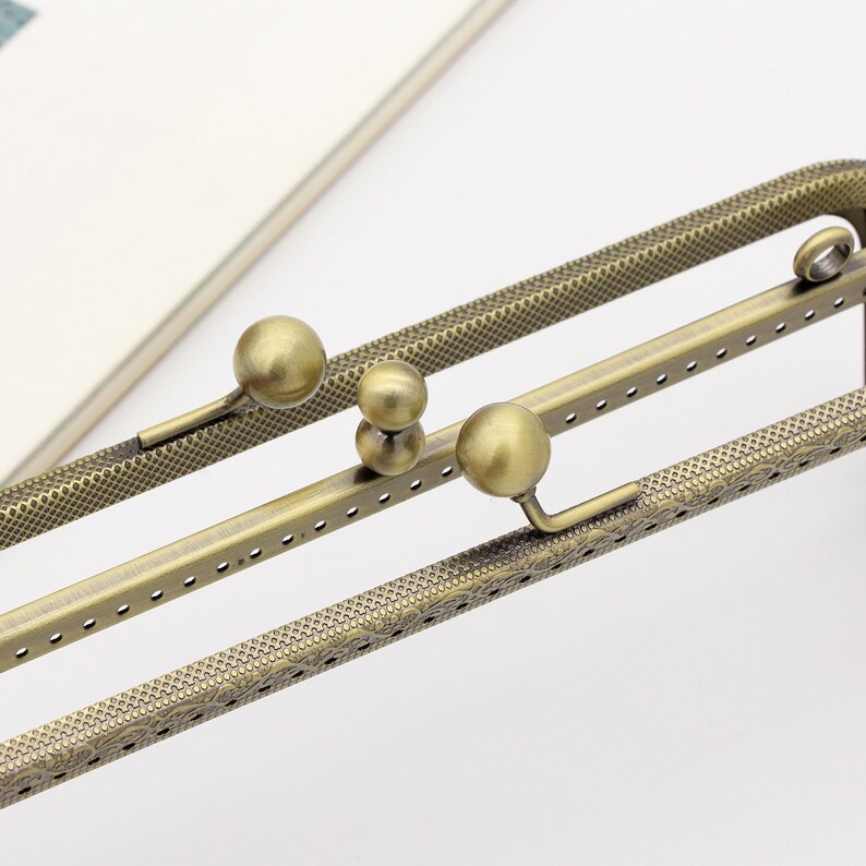 May include: A close-up of a gold-toned metal frame with a decorative pattern. The frame has multiple holes and is designed to be used as a purse or bag closure. The frame has three round metal knobs that can be used to adjust the length of the frame.