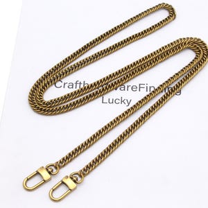 K&ouml;nnte beinhalten: Eine goldfarbene Kette mit einem Curb-Link-Design, ausgestattet mit zwei Karabinerverschl&uuml;ssen. Die Kette wird auf einer wei&szlig;en Oberfl&auml;che pr&auml;sentiert, mit dem Text "CraftwareFinding Lucky" sichtbar.
