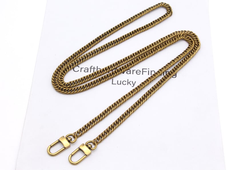 K&ouml;nnte beinhalten: Eine goldfarbene Kette mit einem Curb-Link-Design, ausgestattet mit zwei Karabinerverschl&uuml;ssen. Die Kette wird auf einer wei&szlig;en Oberfl&auml;che pr&auml;sentiert, mit dem Text "CraftwareFinding Lucky" sichtbar.