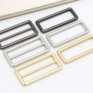 May include: Four metal slider buckles in different finishes: black, silver, gold, and brass. The buckles are rectangular with a double-bar design.