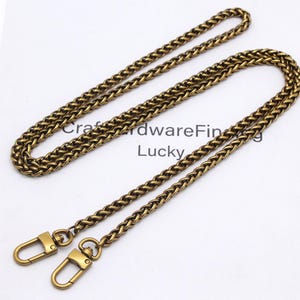 K&ouml;nnte beinhalten: Ein antiker, messingfarbener Kettenriemen mit Karabinerhaken. Die Kette hat ein gewebtes Design und wird auf einer wei&szlig;en Oberfl&auml;che pr&auml;sentiert. Der Text "Craft Hardware Finding Lucky" ist im Hintergrund sichtbar.